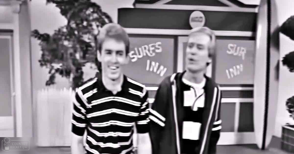 Two young adults in a casual indoor setting, possibly a hotel lobby; one wearing a striped polo shirt and the other a varsity jacket, with vintage signs reading 'Surf Inn' visible in the background.