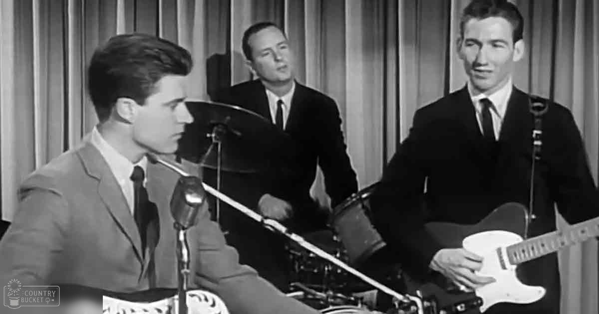 Young adult male singers in a mid-20th century musical performance scene with dark suits and light shirts under collared shirts, set against a simple curtain backdrop.