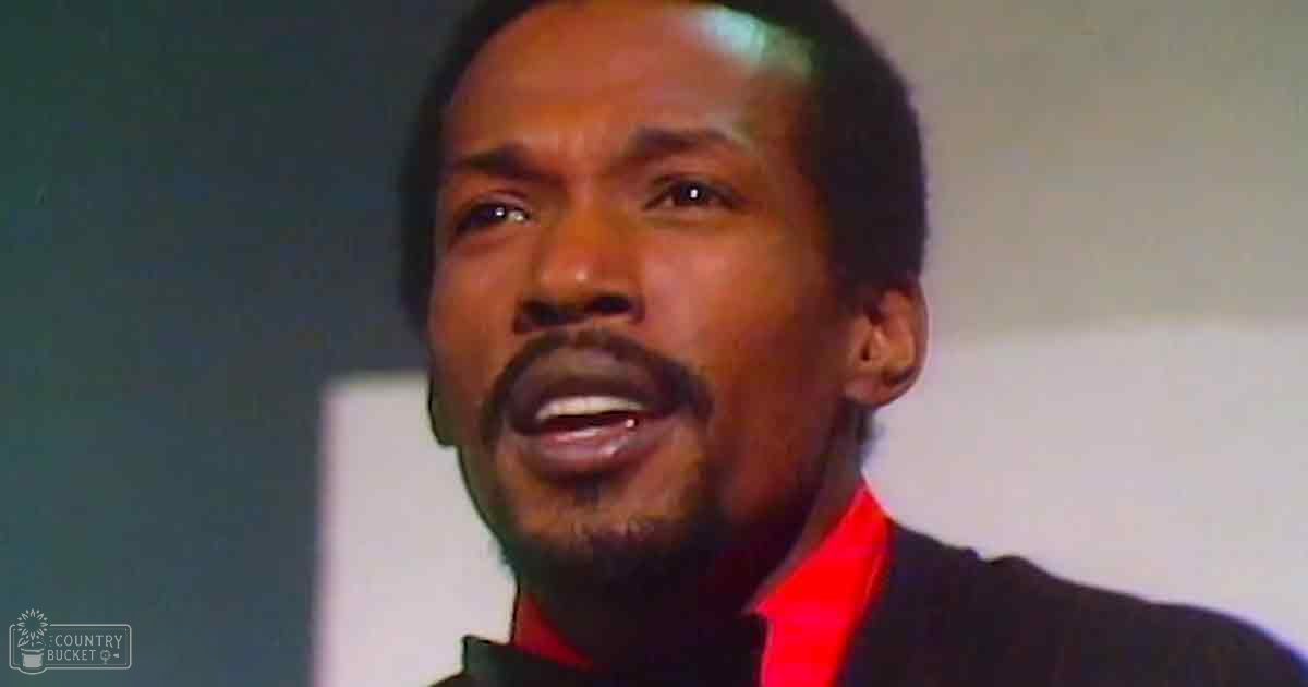 Middle-aged adult male wearing a formal dark-colored suit jacket with a red shirt underneath, set against an indoor plain background.