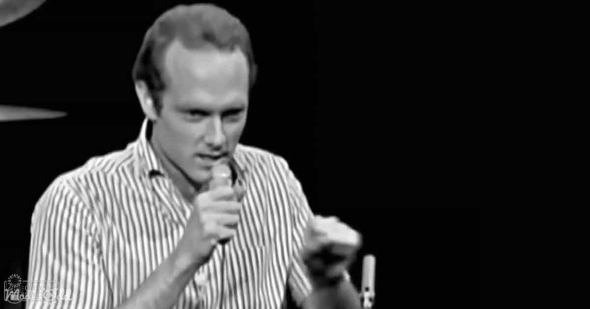 A middle-aged man wearing a collared, button-down, striped short-sleeved shirt with vertical black stripes against a dark indoor background, possibly part of a stage performance or presentation.
