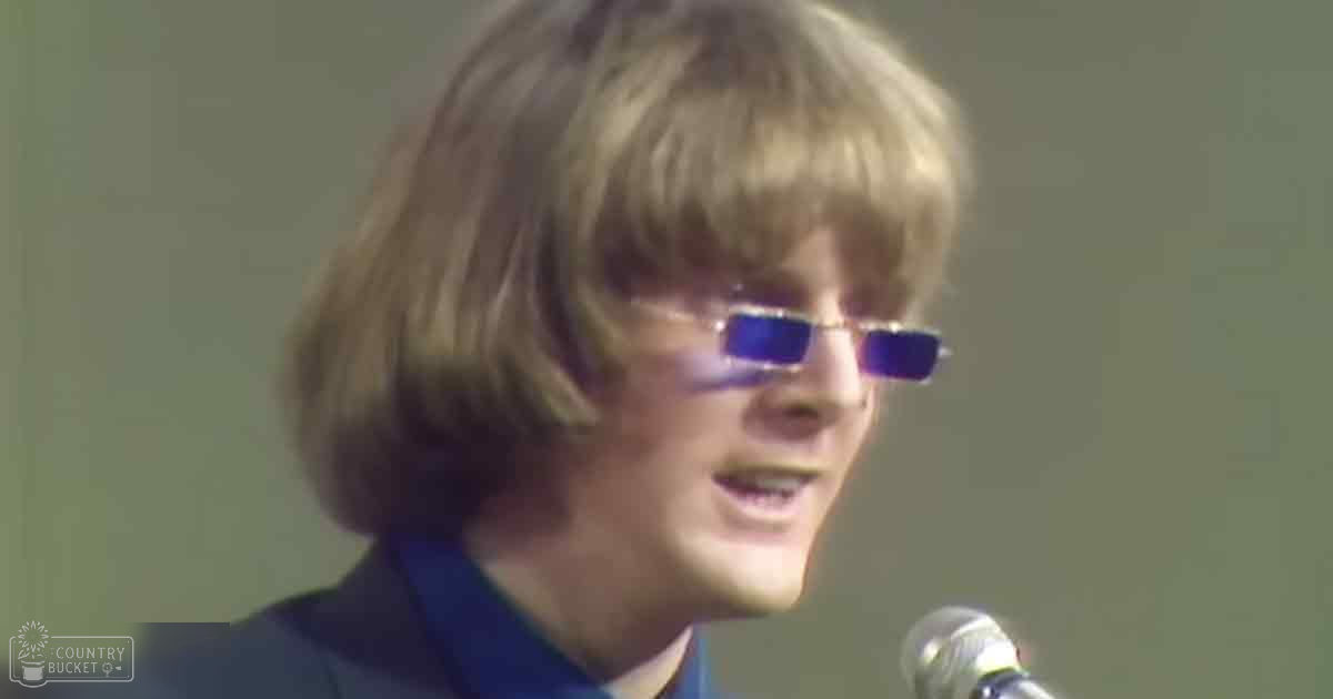 Young adult male wearing a dark-colored, form-fitting jacket with high collar and long sleeves, standing in an indoor setting with a microphone in front of him.