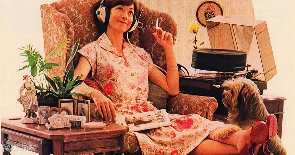 A middle-aged adult female in a floral dress indoors with a small brown and white Shih Tzu dog. The setting appears to be a living room, possibly spring or summer based on the attire and indoor plants.