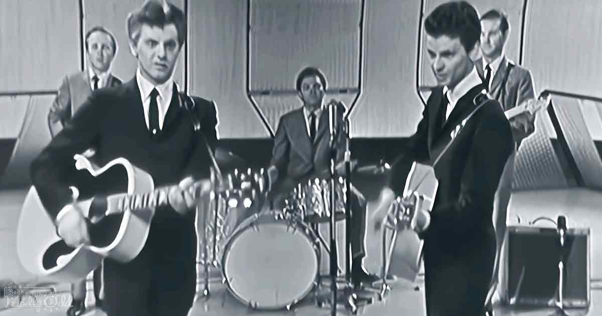 Young adult male musicians in formal attire perform indoors, with a drum set and microphones in focus against wooden-paneled walls under artificial lighting.