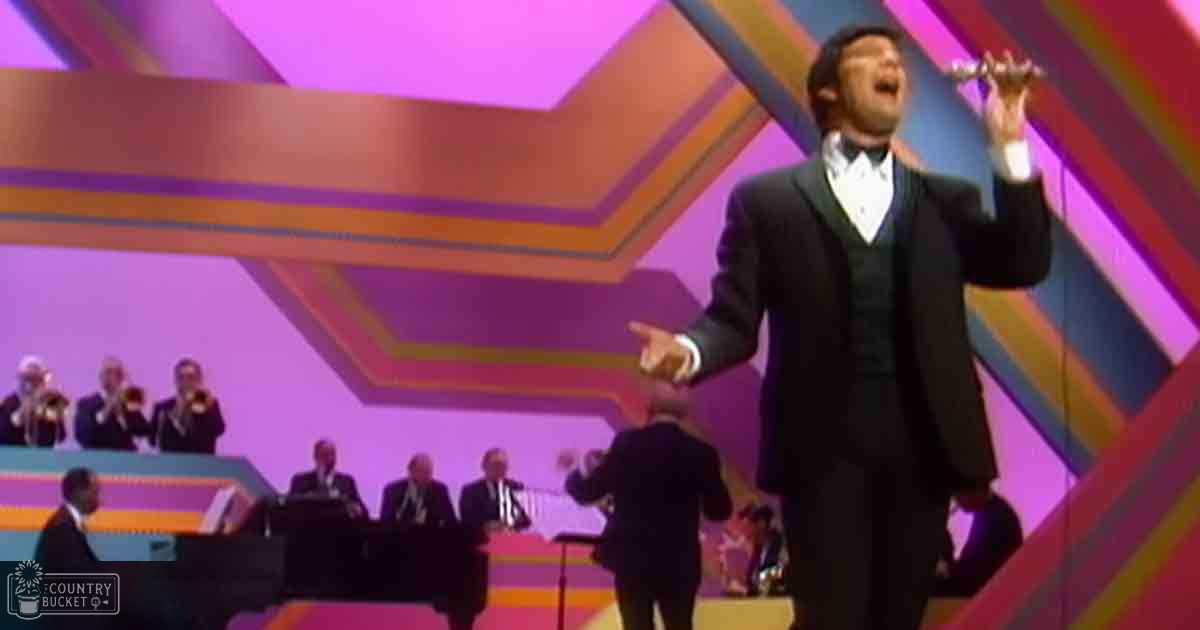 An adult male singer in a black tuxedo performs on stage with musicians dressed in formal suits from the 1970s. The vibrant geometric backdrop and stage lighting create an engaging concert atmosphere.