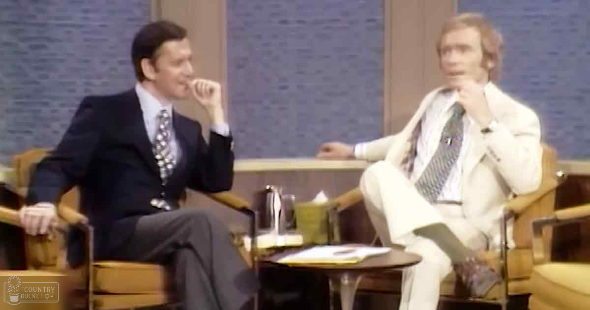 Two adults seated in a talk show setting, one wearing a dark suit jacket and patterned tie, the other in a light-colored suit with reddish-brown hair; textured blue-gray wall suggests indoor studio environment.