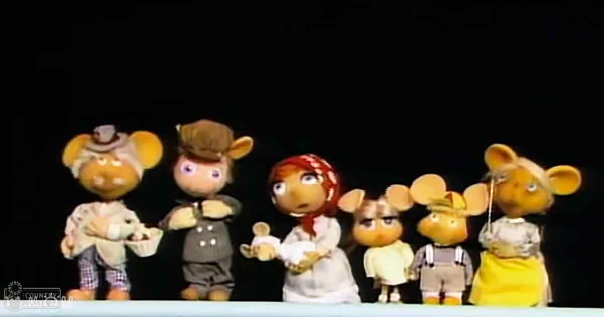 A group of puppet characters in an indoor setting, with a child or young adult female in a light-colored shirt and checkered pants, an adult male in a dark suit jacket and hat, another child or young adult female in a white dress with a red headband, an adult male in a light-colored shirt and dark pants, and an adult female in a yellow dress with a white apron. The background is entirely black.
