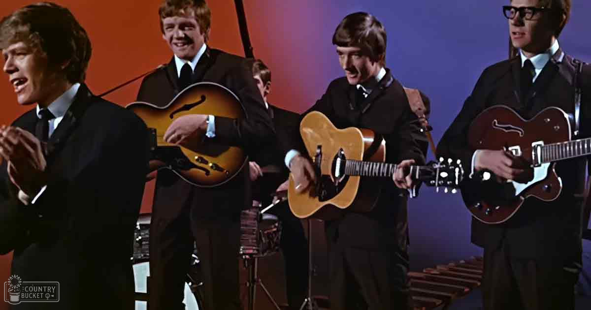 A musical performance scene with young adult males dressed in dark suits on stage under orange and purple lighting.