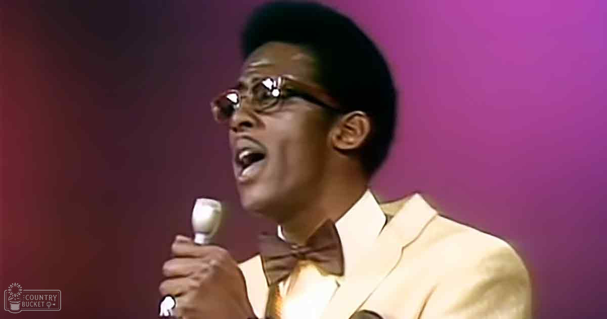 Adult male singer in a studio setting with a light-colored suit jacket and bow tie, holding a microphone.