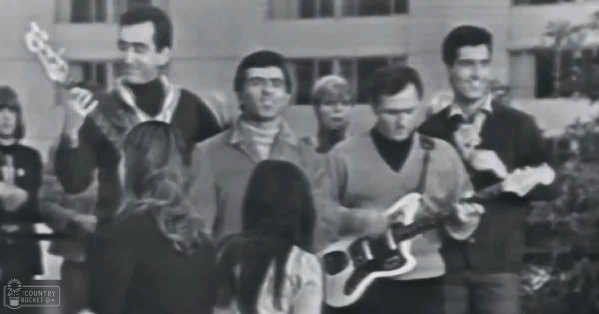 A musical performance in an outdoor setting with people wearing dark jackets and high collars against a backdrop of trees and buildings.