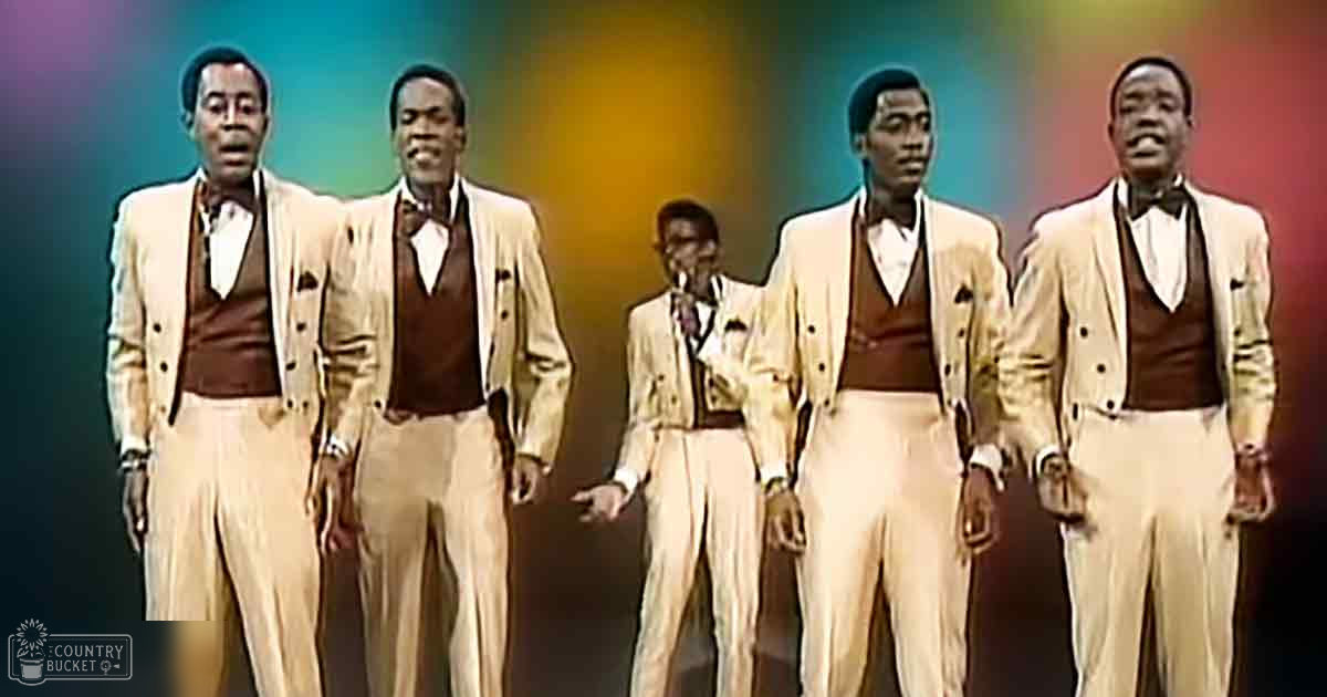 A group of young adult male performers dressed in formal beige double-breasted suits, set against a vibrant purple, green, yellow, blue, and red gradient background.
