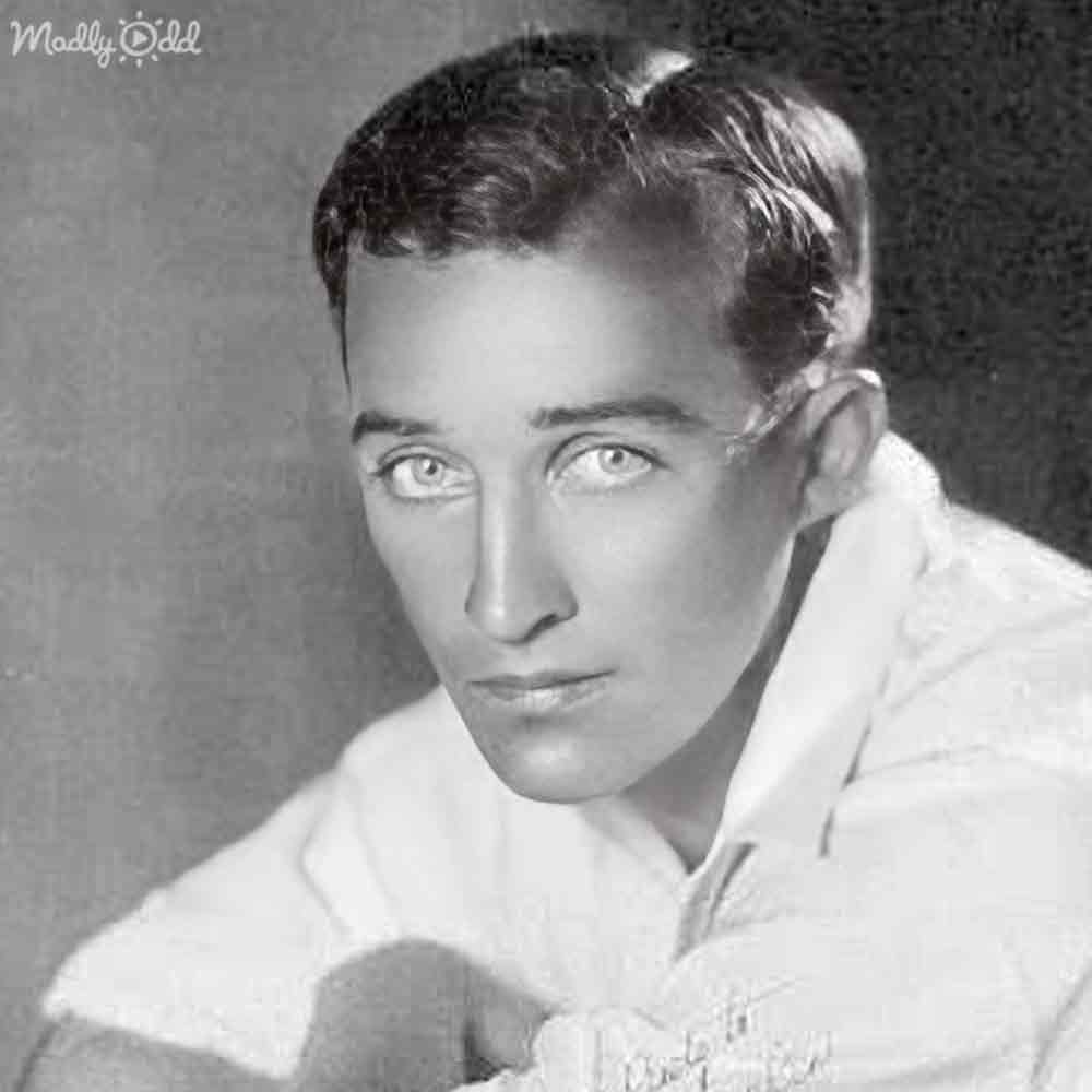 A young adult male wearing a relaxed, early-to-mid-20th-century white shirt with short sleeves and a collar stands indoors against a plain background.
