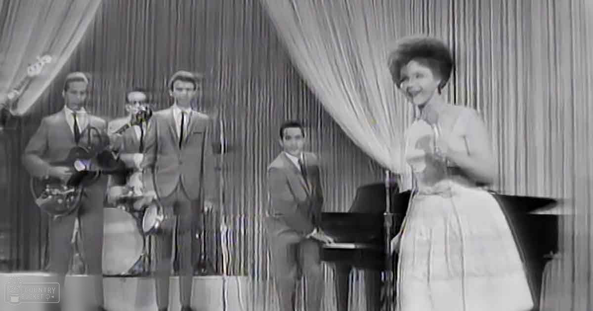 A live musical performance with a woman singing on stage and band members playing instruments under heavy curtains in an indoor setting.