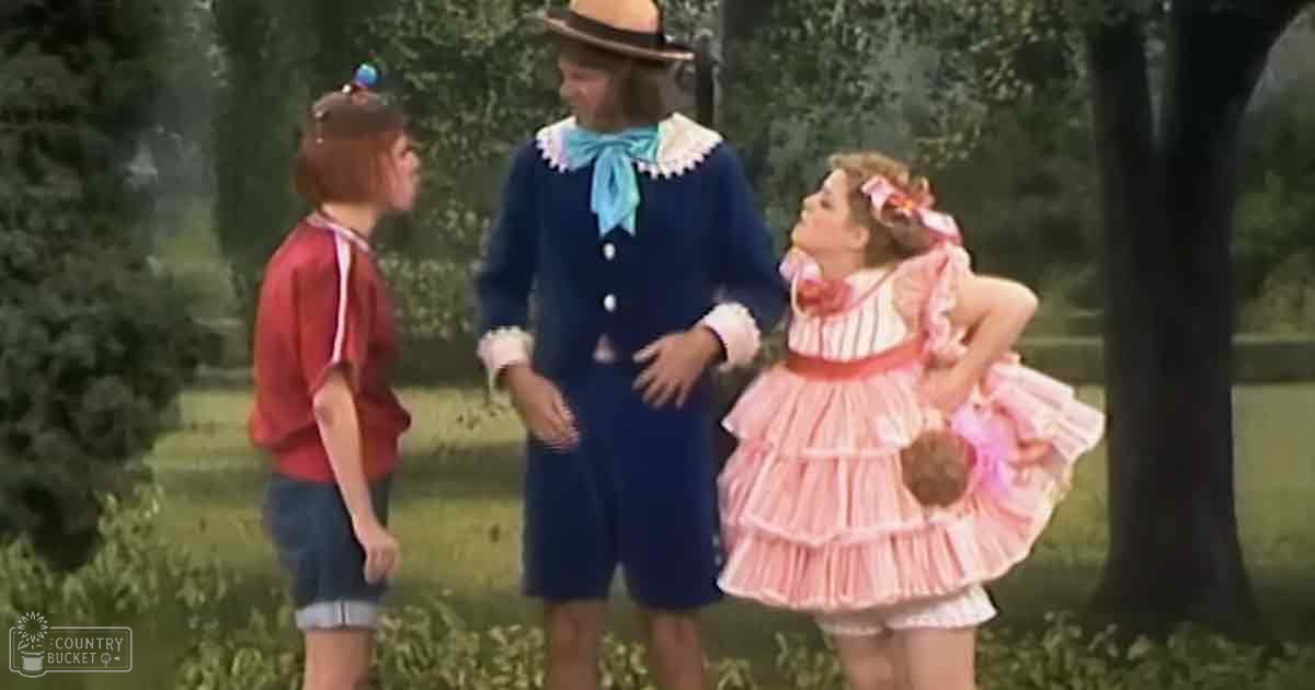 A group of children in colorful outdoor attire, including a child in a pink dress with ruffles and another in a blue romper on a sunny day with greenery in the background.