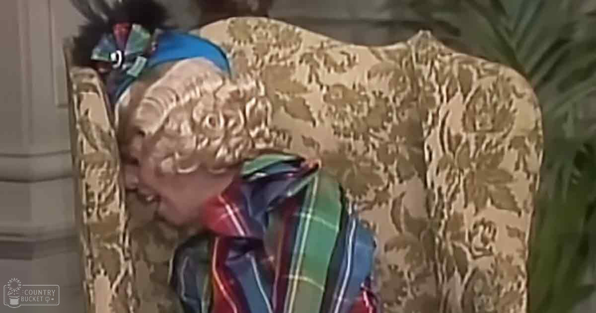 An older adult male wearing a high-collared, relaxed-fit red, blue, green plaid shirt with long sleeves, adorned with a colorful feathered headband, is seated in a patterned armchair with floral upholstery and ornate vintage-style decor. The setting appears to be an indoor living room or similar space within a home.