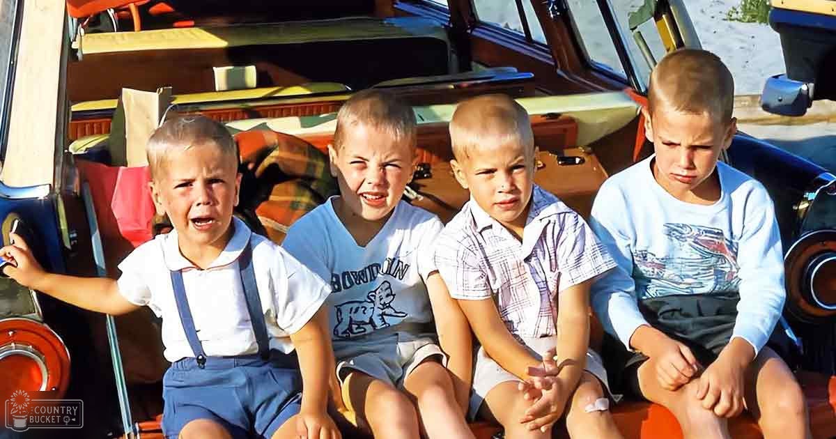 Group of children wearing casual summer clothing sitting in front of an old car at a roadside attraction on a sunny day with clear weather.