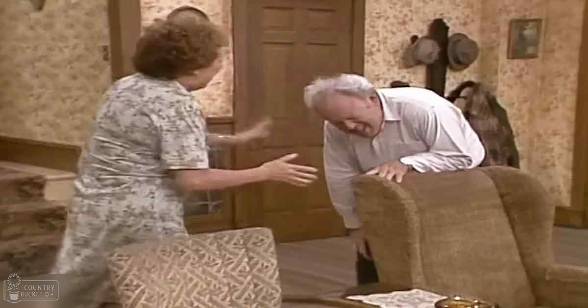 An older adult woman in a light-colored patterned dress interacts with a younger adult man wearing a loose-fitting white shirt, seated in a cozy living room setting with beige armchairs and subtle wallpaper.