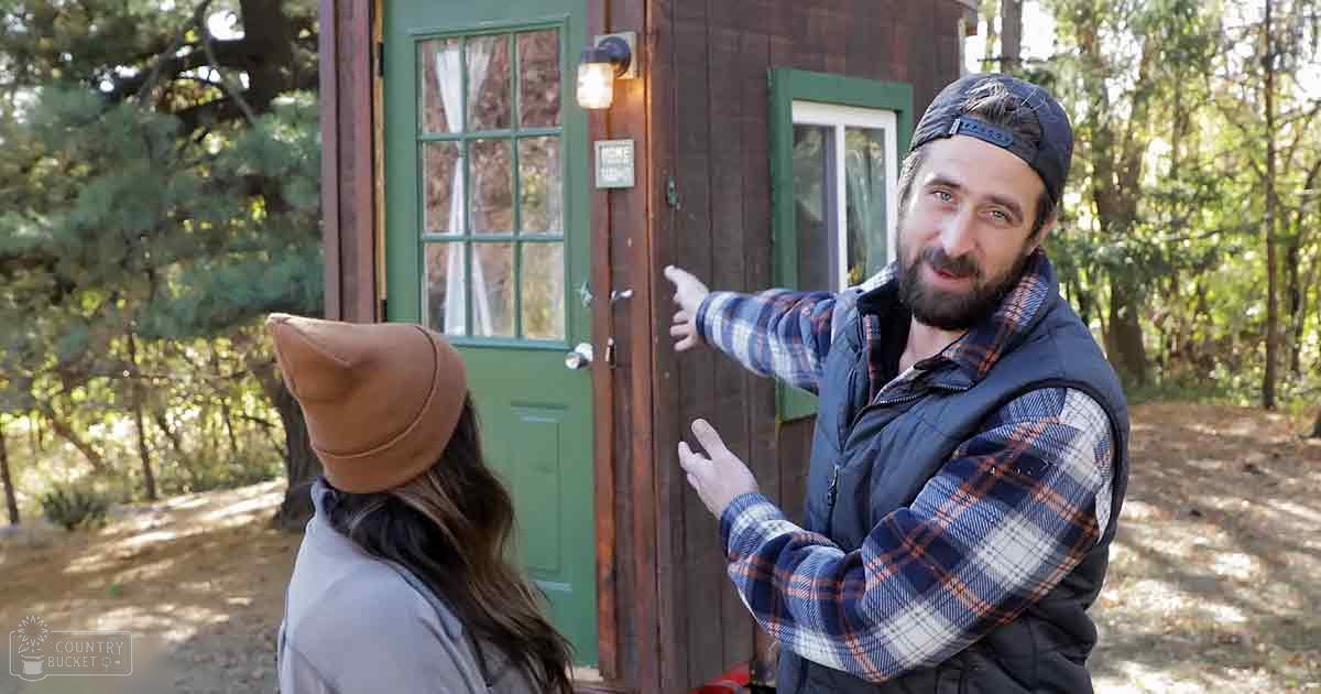 Middle-aged man wearing a blue and white plaid shirt, dark vest, backward cap, and beard, standing outside a small cabin with green-framed windows in a forested area during late summer or early fall.