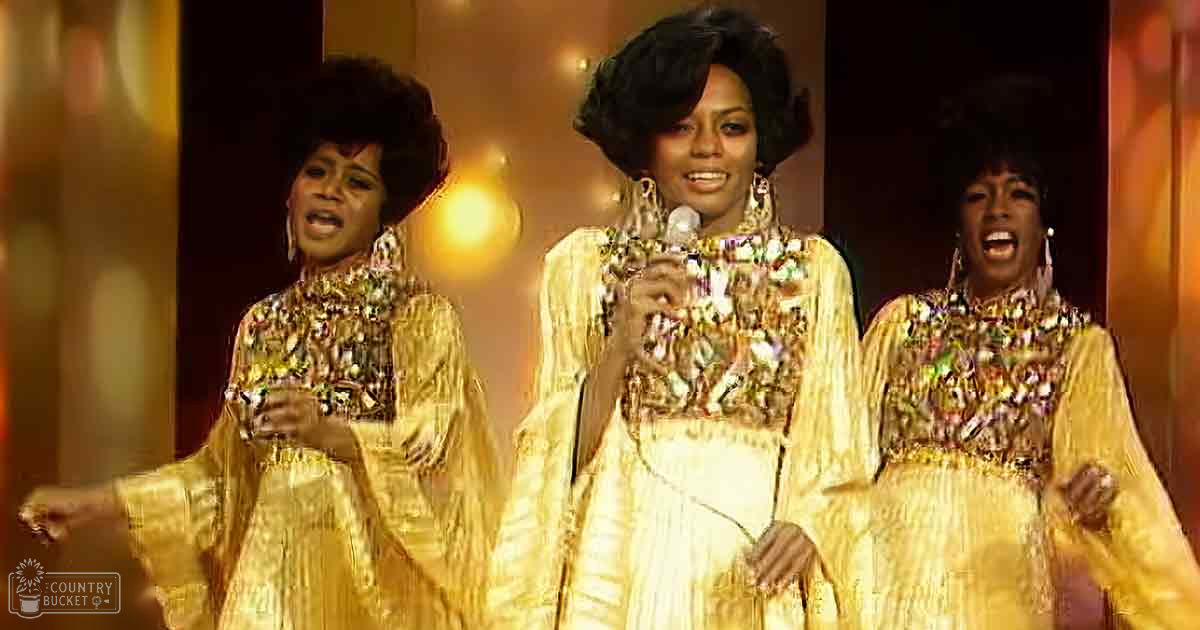 A stage performance scene with three adult females wearing matching gold dresses with intricate patterns and high necklines from the mid-20th century, possibly 1960s, set against a warm-lit indoor venue.