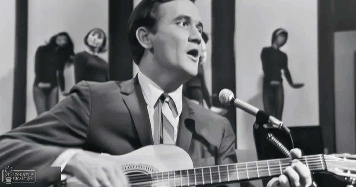 An adult male singer wearing a dark suit jacket with a light-colored tie, holding an acoustic guitar and singing into a microphone in what appears to be an indoor recording studio or performance venue.