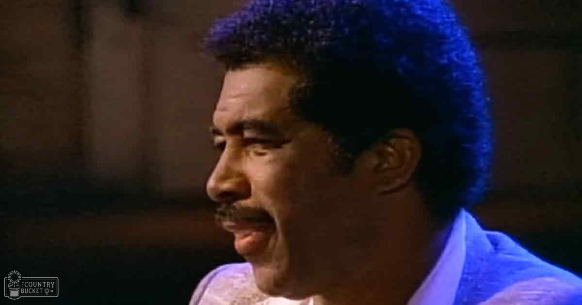 An adult male wearing a mid-20th-century style light blue garment with high collar and short sleeves, seen against an indoor setting with artificial lighting.