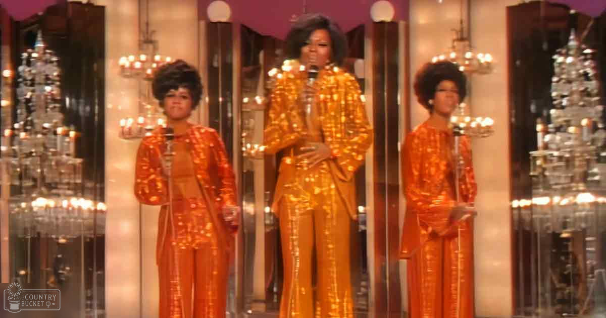 Three adult female performers in shiny orange sequined jumpsuits on a stage with reflective gold from chandeliers and cityscape reflections.
