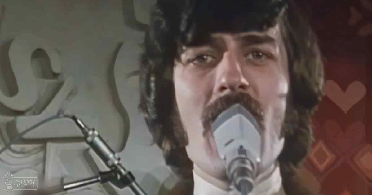 A middle-aged male singer in a recording studio or performance setting, wearing a white shirt with a high collar and long sleeves, singing into a microphone.