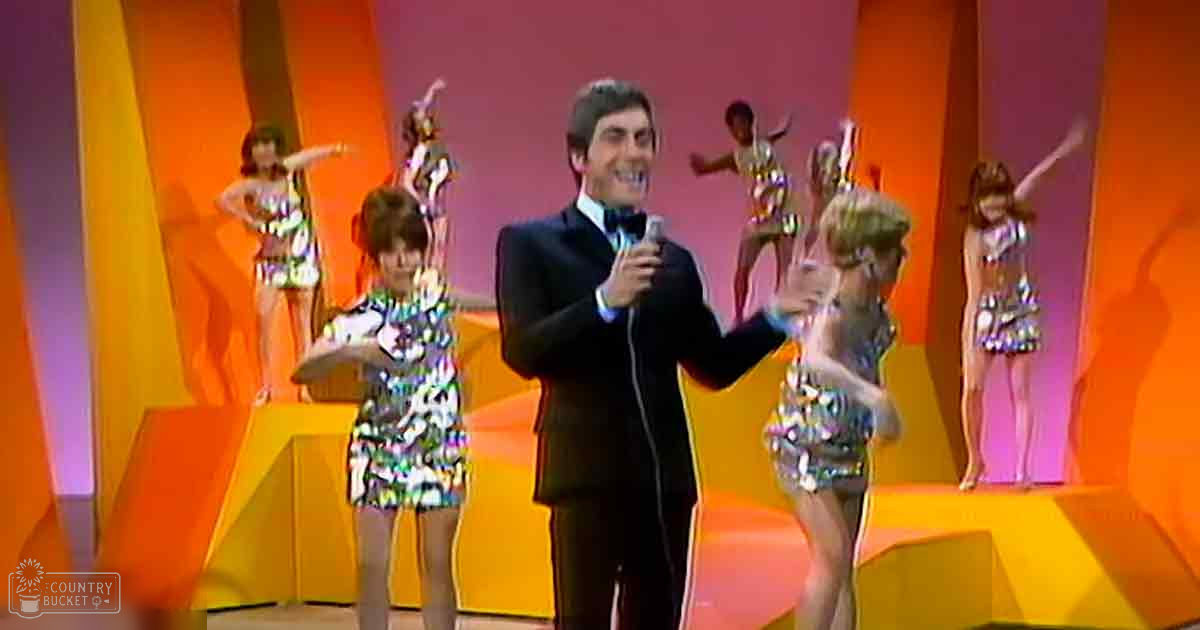 An adult male performer holding a microphone on stage in front of dancers wearing colorful sequined dresses.