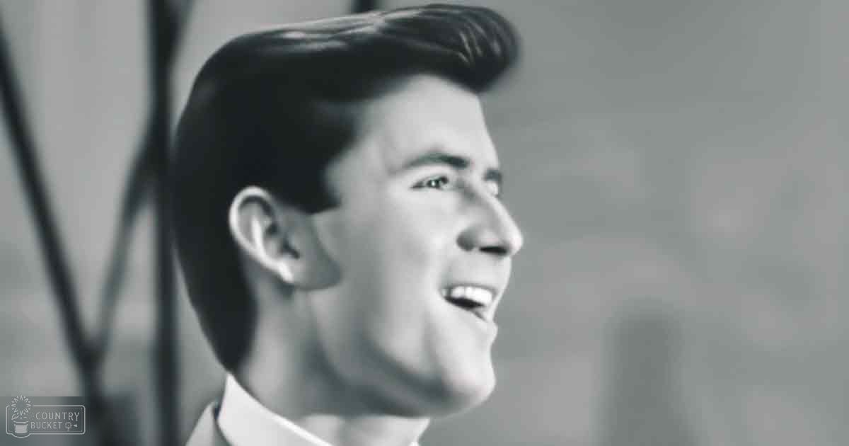 A young adult male in a collared shirt stands against an indoor backdrop with neutral tones and a microphone stand, suggesting a musical performance setting.