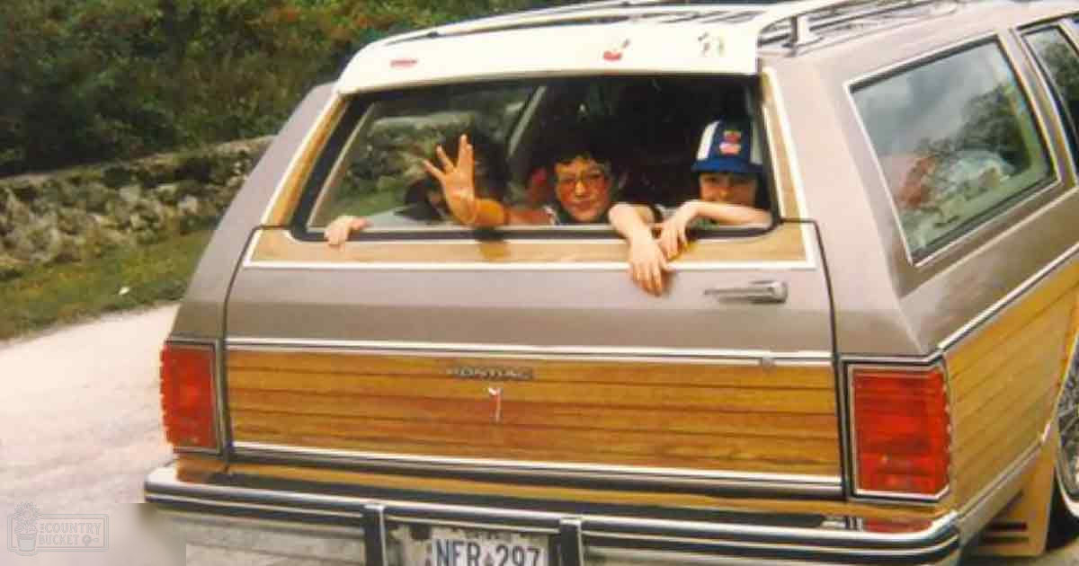 Two children lean out of the rear window of a vintage station wagon in a lush rural area on what appears to be a sunny day.