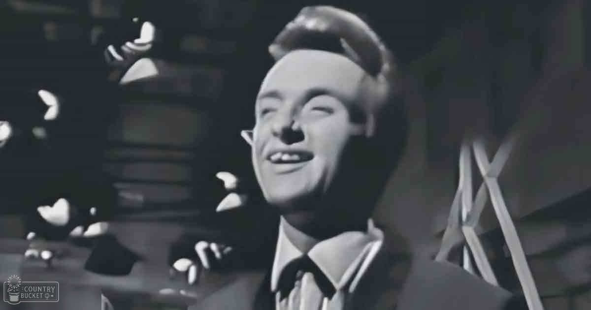 A young adult male in formal attire, possibly a performance or recording session setting with musical instruments and dim stage lighting.