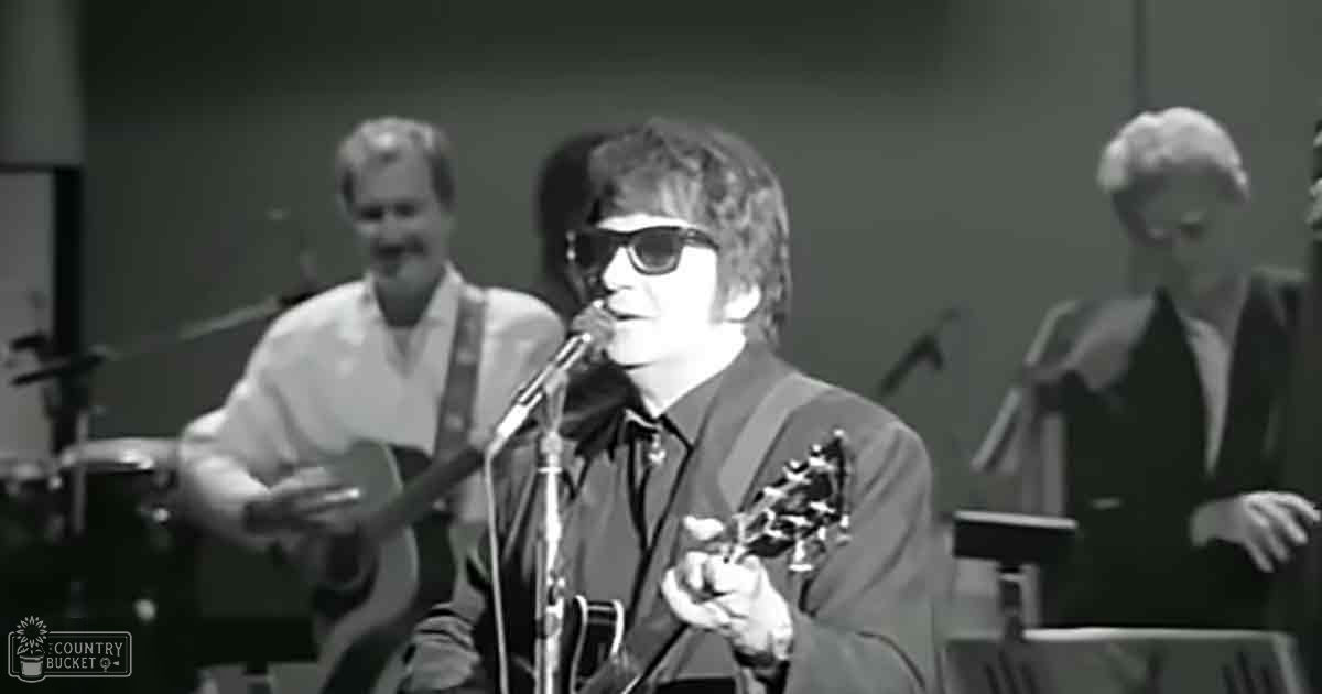 An adult male performer in a dark suit jacket and shirt singing into a microphone while playing guitar, with other musicians in an indoor performance venue.