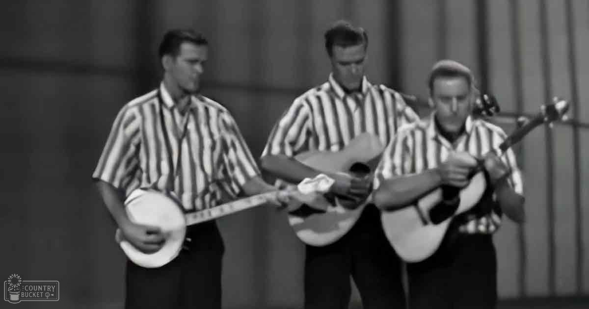 Young adult males performing in a musical scene with striped shirts, set against an indoor backdrop resembling curtains or blinds under even lighting.