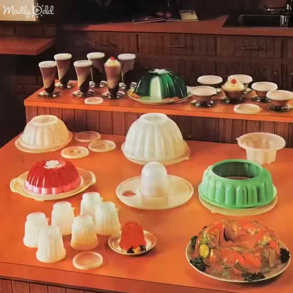 Vintage plastic jello molds and colorful layered desserts displayed indoors, possibly a kitchen or dining area with wooden cabinets and countertops.