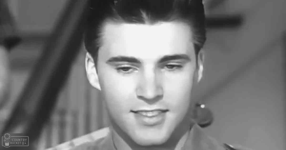 A young adult male wearing a mid-20th-century plaid shirt indoors with curtains and wooden elements in the background.
