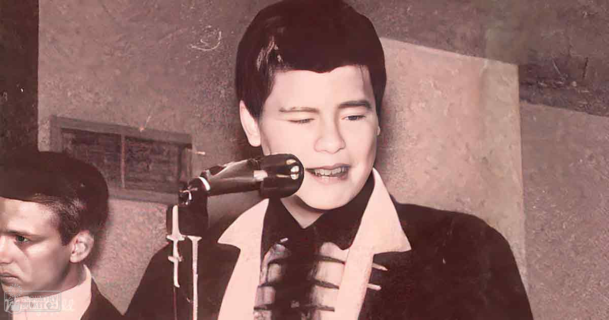 A young adult male singer wearing a dark suit jacket with white lapels and a patterned tie stands indoors near another individual also in formal attire.