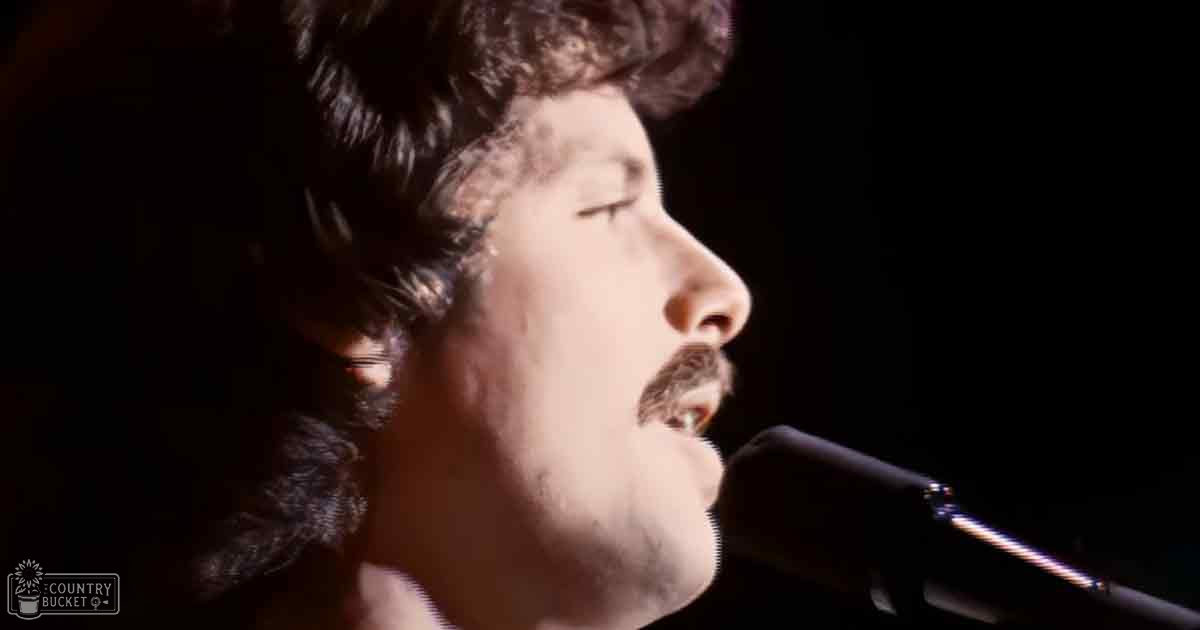 A young adult male singer wearing a dark-colored, fitted garment with long sleeves and smooth material, holding a microphone indoors under controlled lighting.