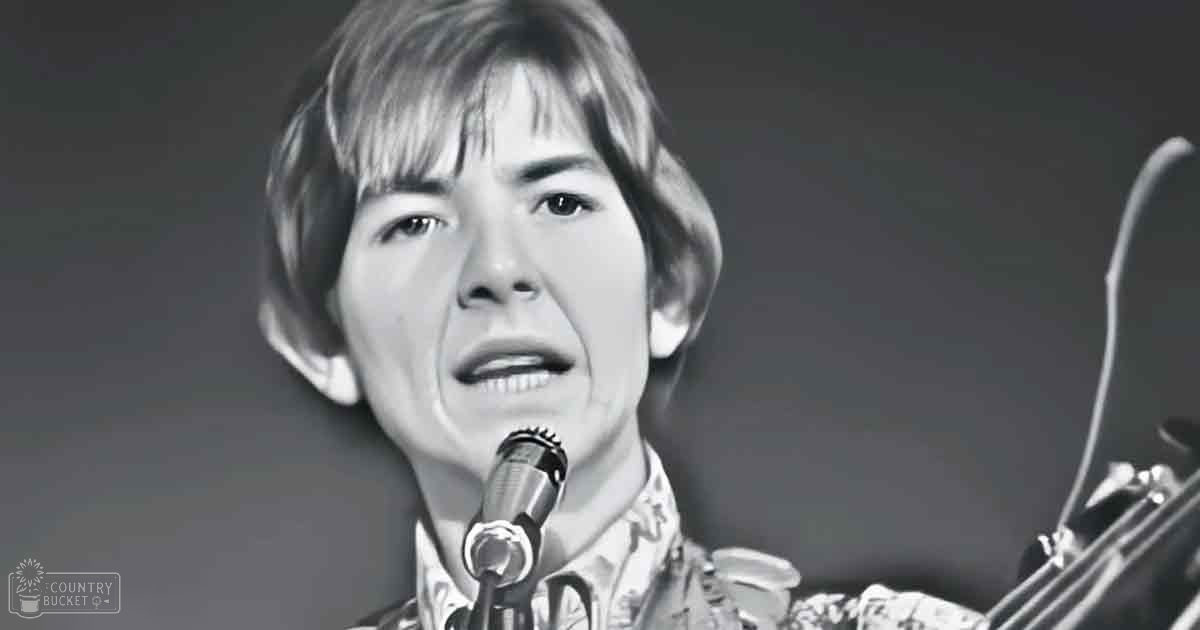 A young adult male singer performs on stage, wearing a patterned shirt with floral or paisley design, likely mid-20th century cotton or linen, in front of a microphone. The setting is an indoor performance space with dramatic stage lighting.