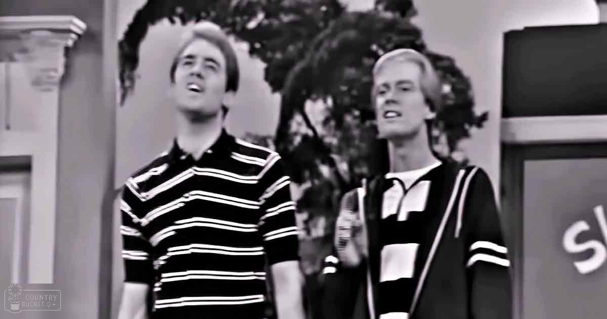 Two young adult males in casual attire, standing indoors with greenery visible outside, wearing striped polo shirts suggesting mid-20th-century style.