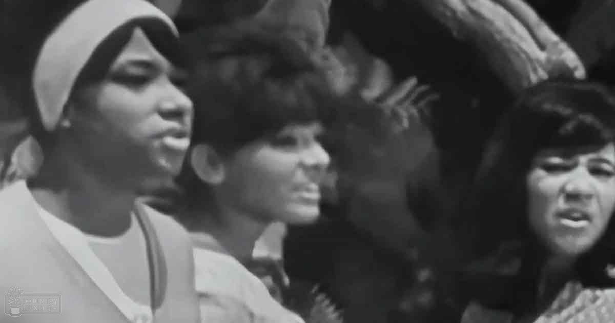 A young adult female wearing a white headscarf and mid-20th century style clothing at an outdoor event with a crowd in the background, possibly a protest or rally.
