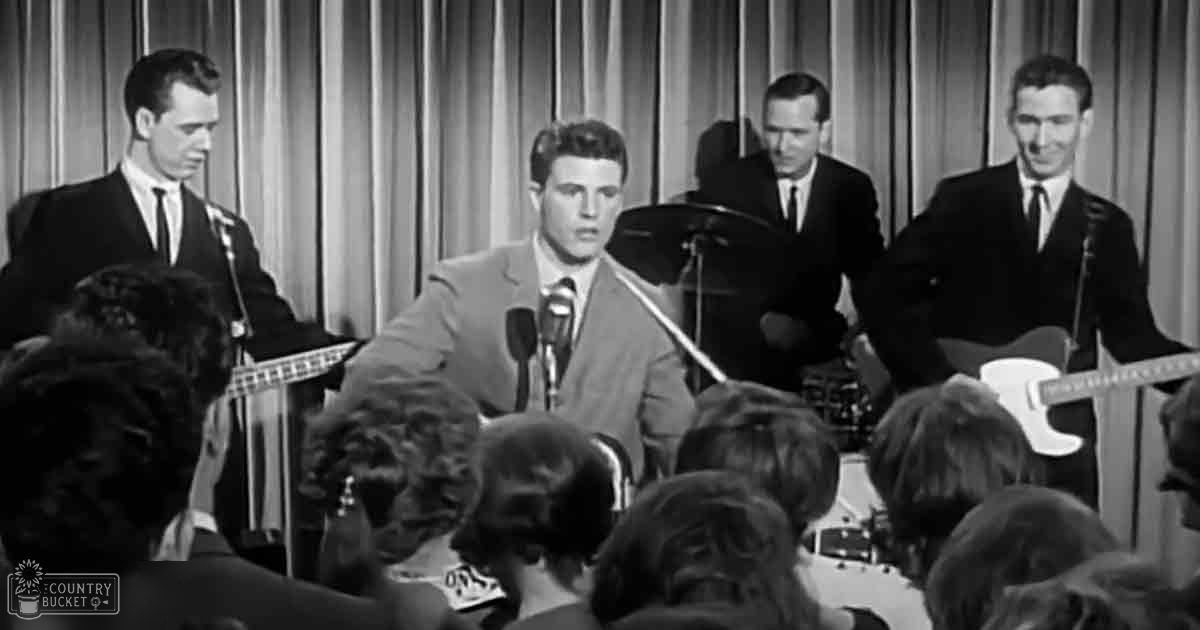 A young adult male singer performing on stage with musicians holding electric instruments at a music venue or concert hall.
