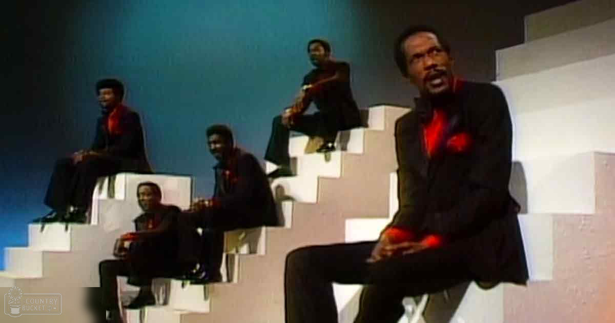 A group of adult males in a staged indoor setting, dressed uniformly in dark suit jackets over red shirts, seated on white steps against a simple backdrop under controlled lighting.