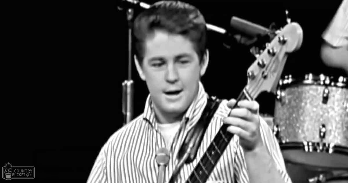 A young adult male playing guitar on a stage in an indoor performance venue with musical instruments and stage lighting.