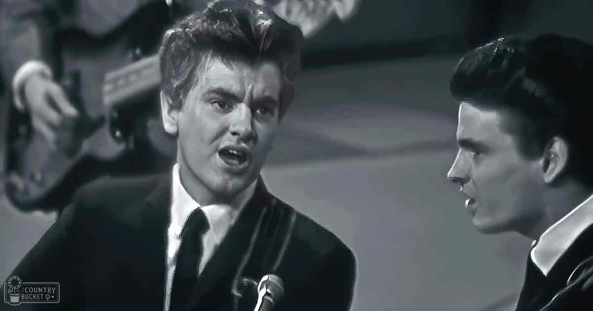 A dimly lit indoor stage with two young adult male performers in dark suits, possibly from a mid-20th century music venue.
