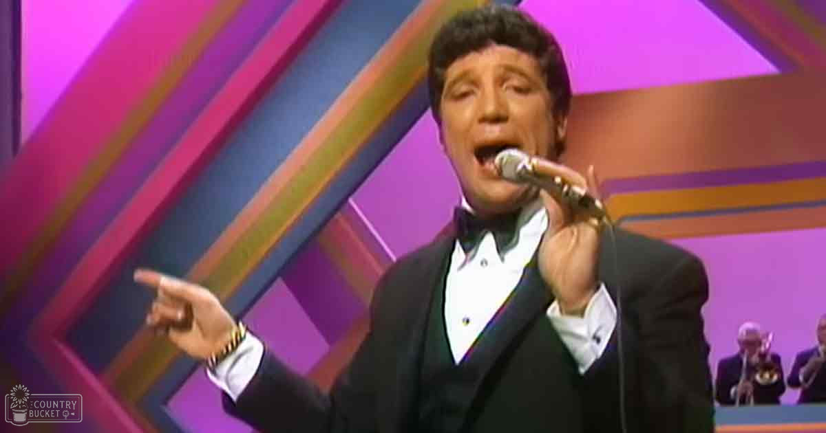 An adult male singer wearing a dark suit with white shirt and black bow tie, performing on stage amidst an audience of middle-aged to older adults in formal attire.
