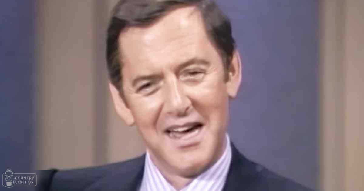 A middle-aged adult male wearing a dark, tailored suit jacket with a striped, light-colored collared shirt under it, in an indoor professional or interview-style setup.