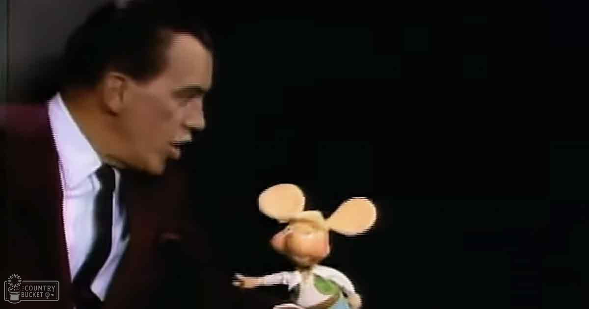 An adult male in a dark suit jacket over a white dress shirt and black tie interacts with an anthropomorphic mouse-like animated character in a studio setting.
