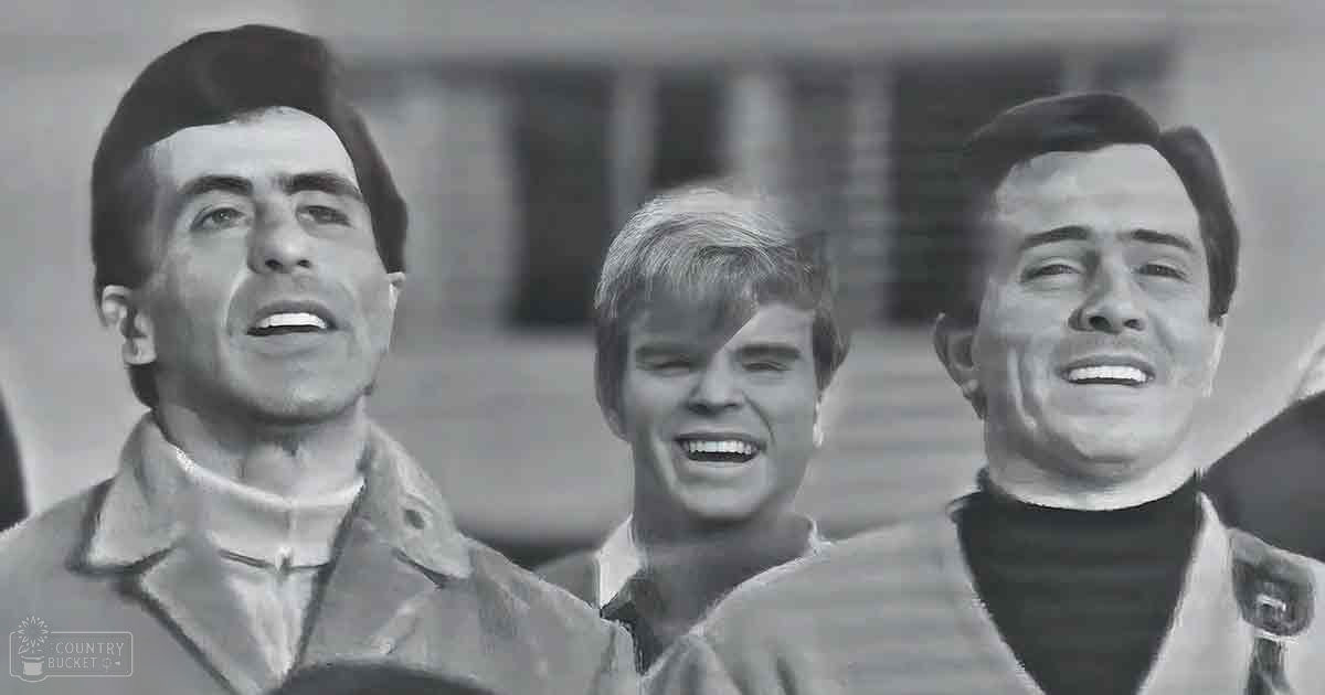 A group of young adult males in a relaxed outdoor setting, wearing jackets with high collars and long sleeves, possibly in cooler weather; architectural elements suggest an urban environment.