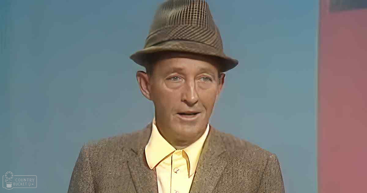 An older adult male in a studio setting, wearing a brownish-gray blazer over a yellow collared shirt with a fedora-style hat, against a simple blue backdrop and red structure.