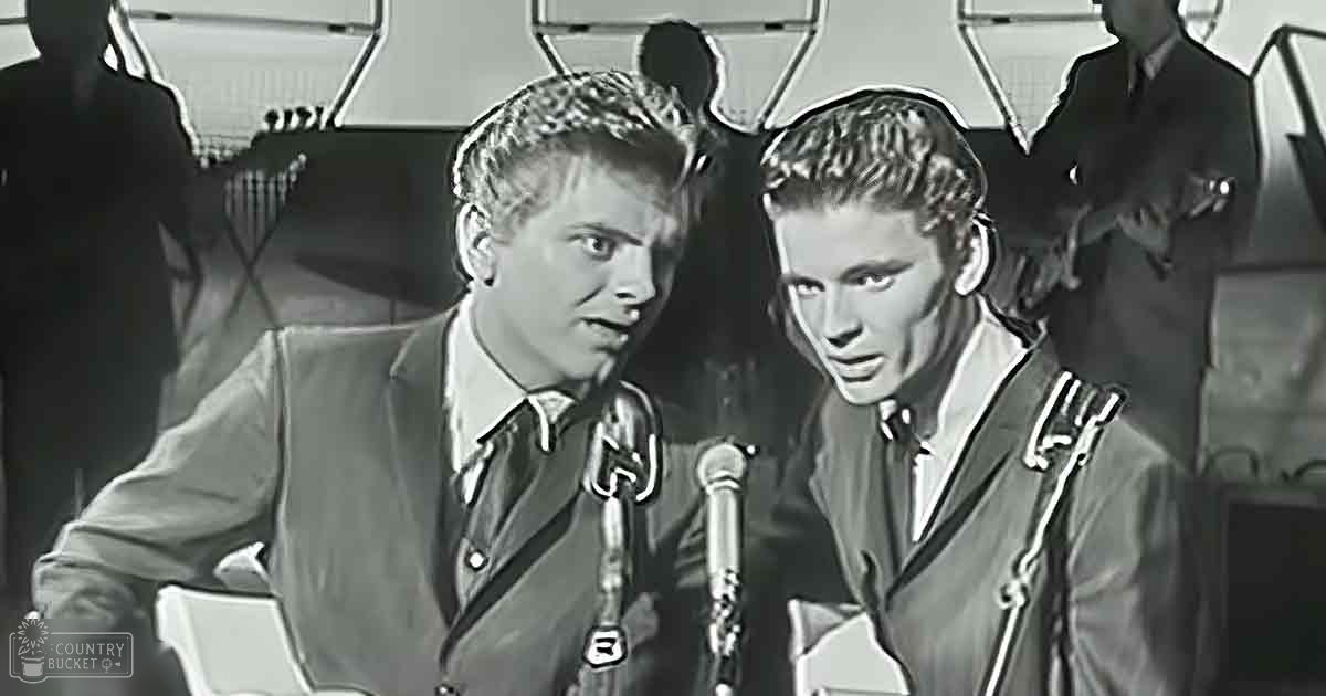 Two young adult males wearing dark-colored tailored suits with white shirts, standing indoors in what appears to be a professional recording studio setting.