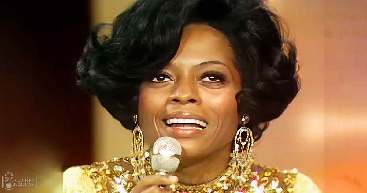 A woman wearing an intricately embellished garment with large ornate earrings, holding a microphone in what appears to be a stage or performance setting.